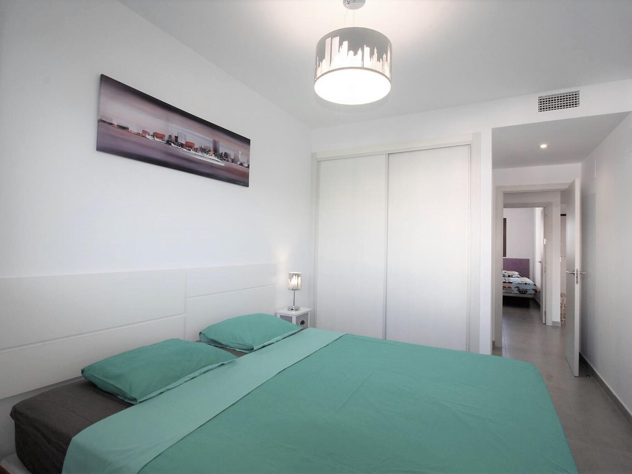 Een slaapkamer met een bed, nachtkastje, lamp, deur en een gang naar een andere kamer.