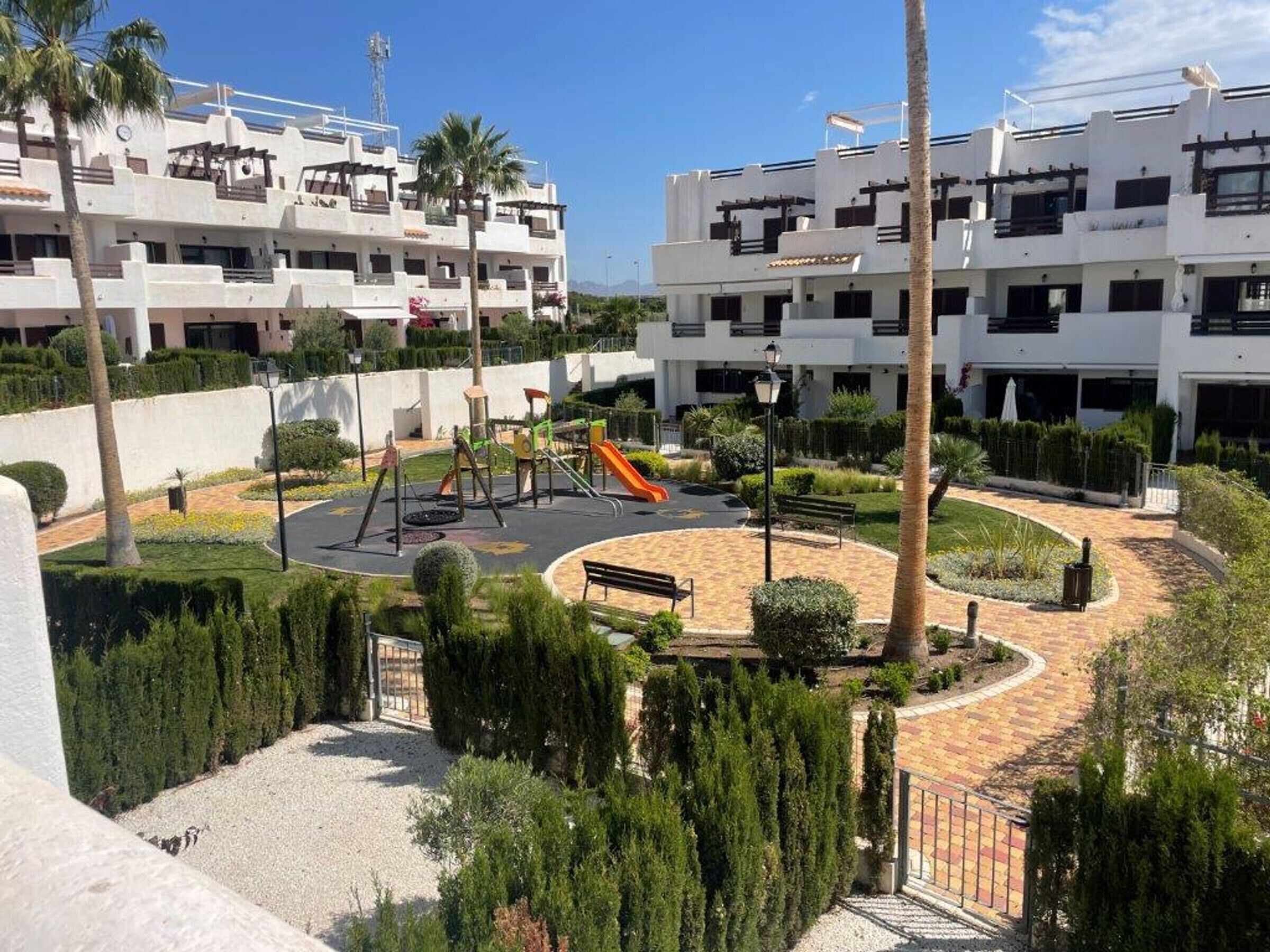 Un parque infantil con columpios y tobogán, rodeado de una zona ajardinada con bancos y palmeras.