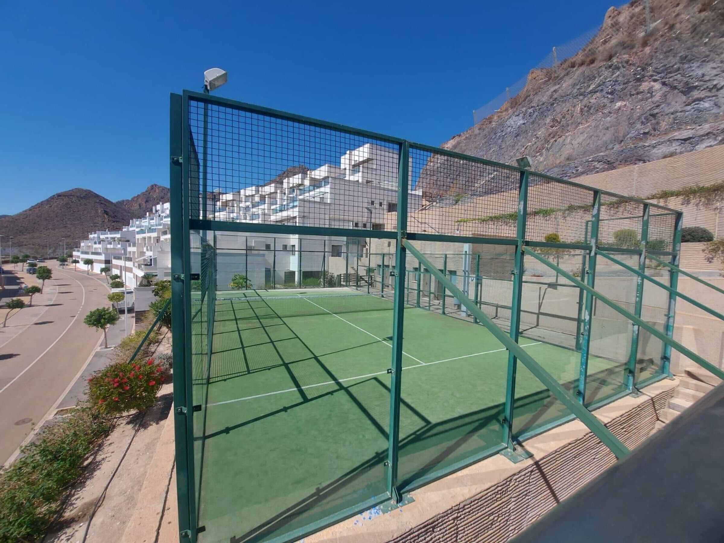 Een tennisbaan, omheind door een groene metalen hekken, met zicht op een woonwijk en een berg op de achtergrond.