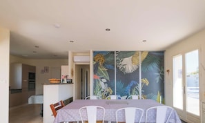 A dining area with a table set for four, a kitchen in the background, and a tropical-themed sliding door. - Cozy Villa in Biscarrosse with Heated Pool, Close to Center, Sleeps 9 (Biscarrosse)