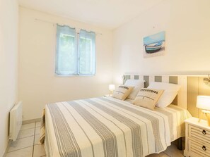 A bedroom with a bed, a window with curtains, a bedside table with a lamp, and a wall-mounted picture. - Charming 3-Bed House in Biscarrosse with Large Terrace and Garden (Biscarrosse)