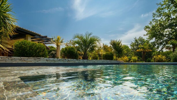 A clear swimming pool with a stone wall and tropical vegetation. - 5-star villa, heated pool, 4 rooms with private baths and BBQ - Up to 12 guests (Parentis-en-Born)