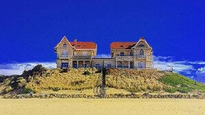 A large, two-story building with a red roof, situated on a sandy beach with a clear blue sky. - Charming 2-bedroom house with garden near the lake, perfect for holidays (Sanguinet)