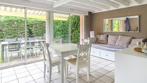 A modern room with a dining table and chairs, a bed with a headboard, and a large window offering a view of greenery. - Biscarrosse: Duplex with Garden, 6 Pers. and Private Parking (Biscarrosse)