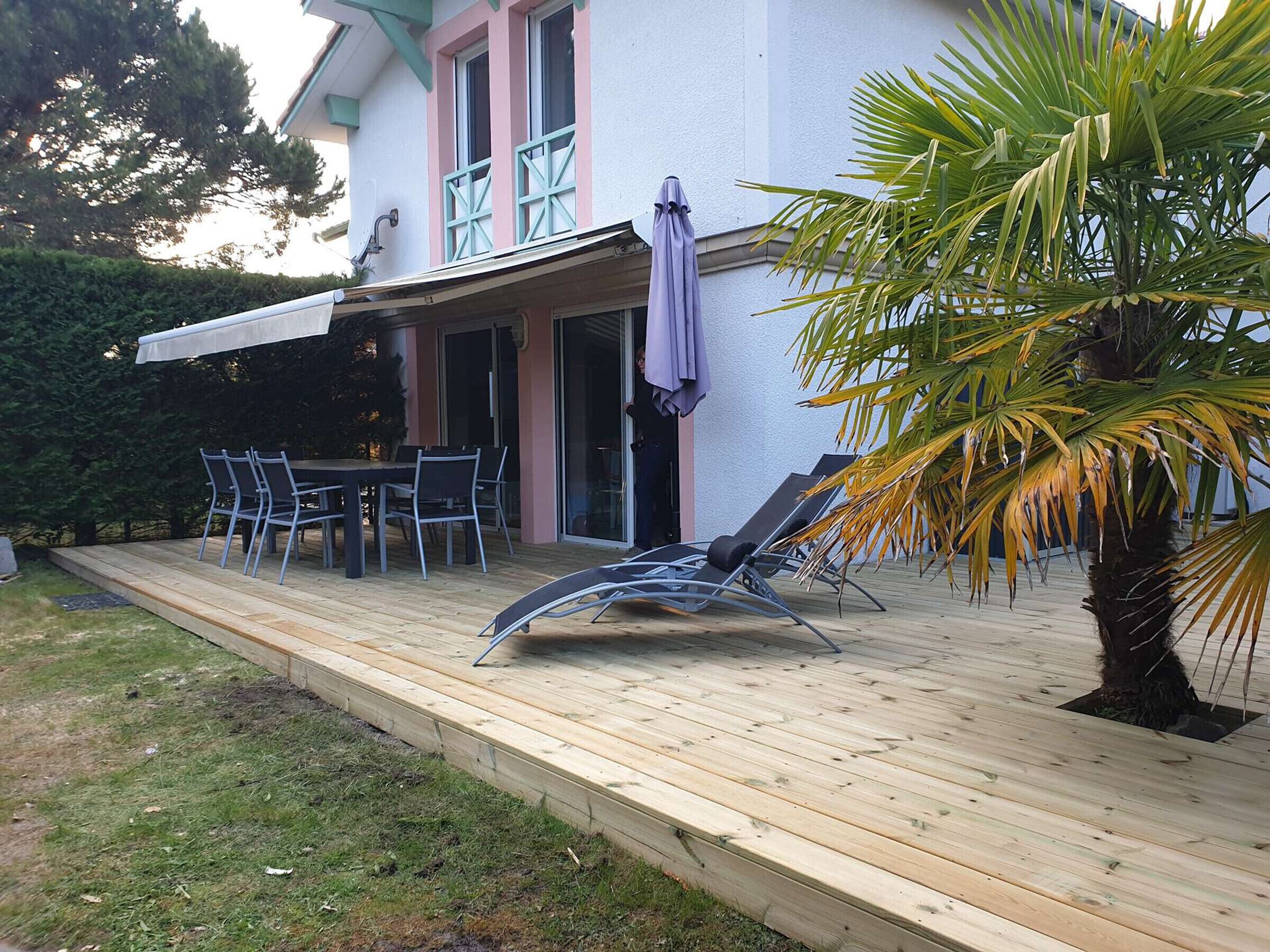 A covered outdoor patio with a table and chairs, a palm tree, and a building with a white exterior and pink trim.