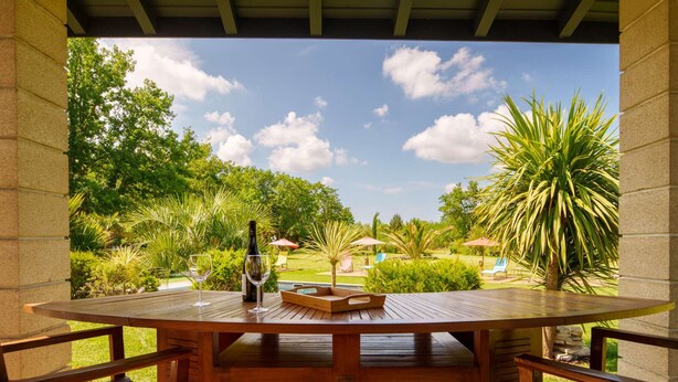 A wooden table with chairs, a bottle, and glasses on a patio with a view of a garden and trees. - 5-Star Villa: Heated Pool, 24 people, near Parentis-en-Born lake (Parentis-en-Born)