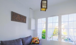 A room with a gray sofa, a colorful pillow, a wall-mounted artwork, and a window with sheer curtains. - Charming 6-Person House with Garden, Terrace, and Outdoor Shower (Sanguinet)