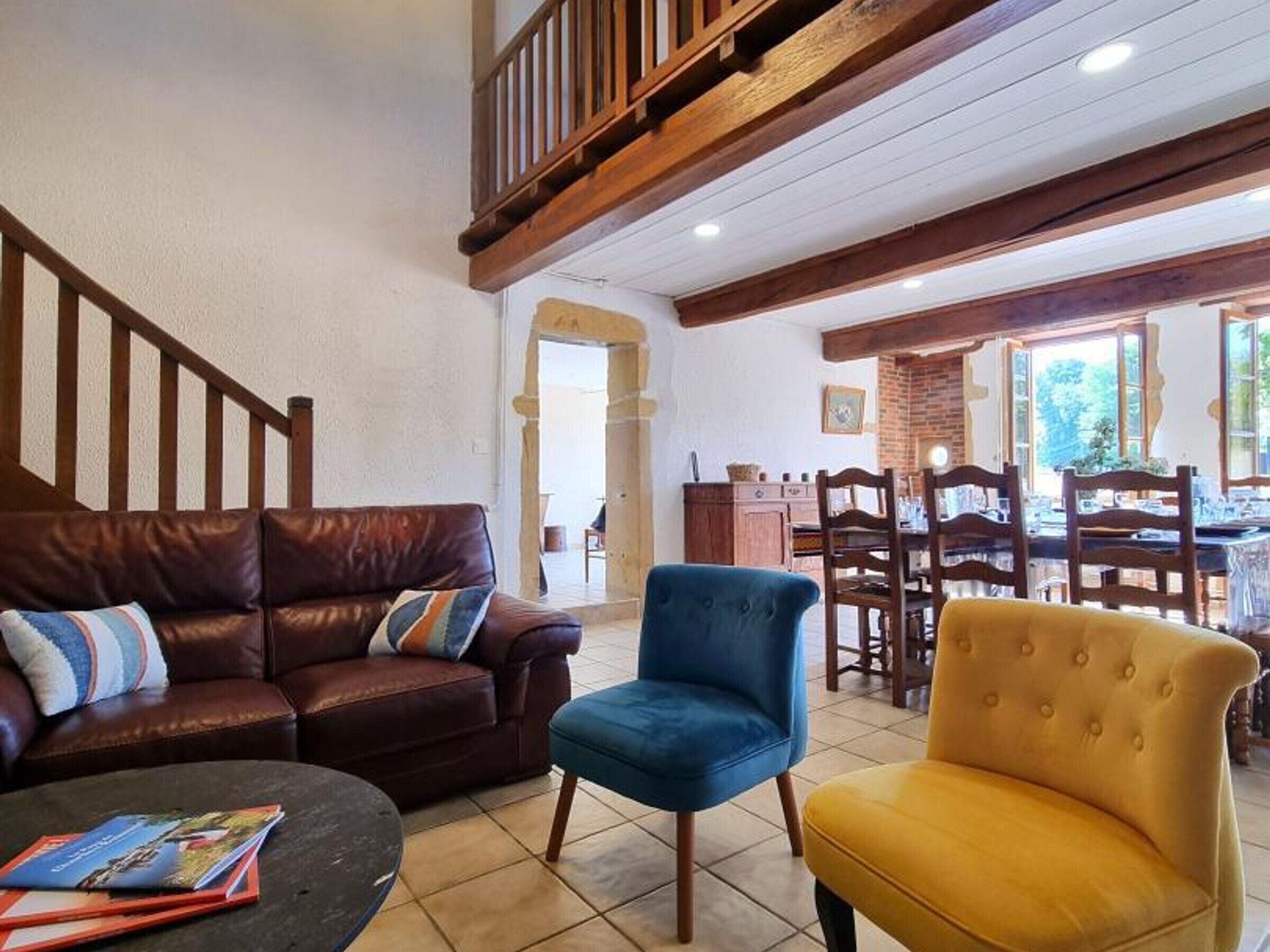 Un salon avec un canapé en cuir marron, un fauteuil bleu et un fauteuil jaune. On y trouve un escalier avec des rampes en bois, un coin repas avec des chaises et une table en bois, ainsi qu’une cheminée en briques.