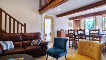 Un salon avec un canapé en cuir marron, un fauteuil bleu et un fauteuil jaune. On y trouve un escalier avec des rampes en bois, un coin repas avec des chaises et une table en bois, ainsi qu’une cheminée en briques.