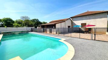 Une piscine à bord incurvé, un espace patio aménagé avec du mobilier d’extérieur et un bâtiment doté d’un toit en tuiles.
