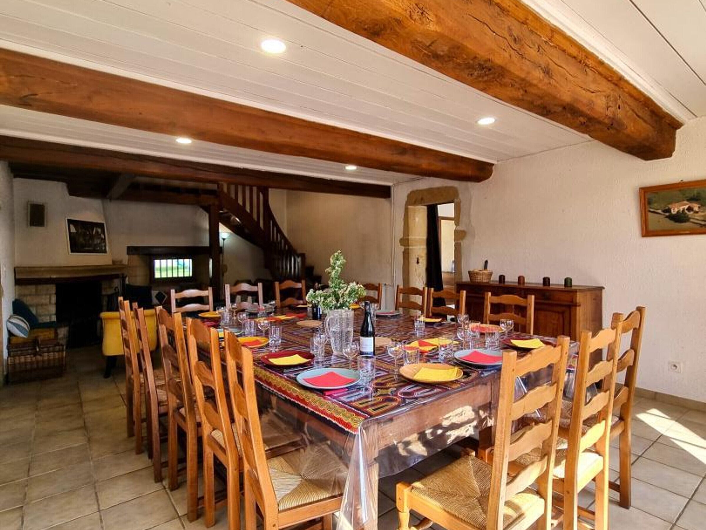 A dining area with a long table set for multiple people, wooden chairs, a chandelier, and a staircase in the background.