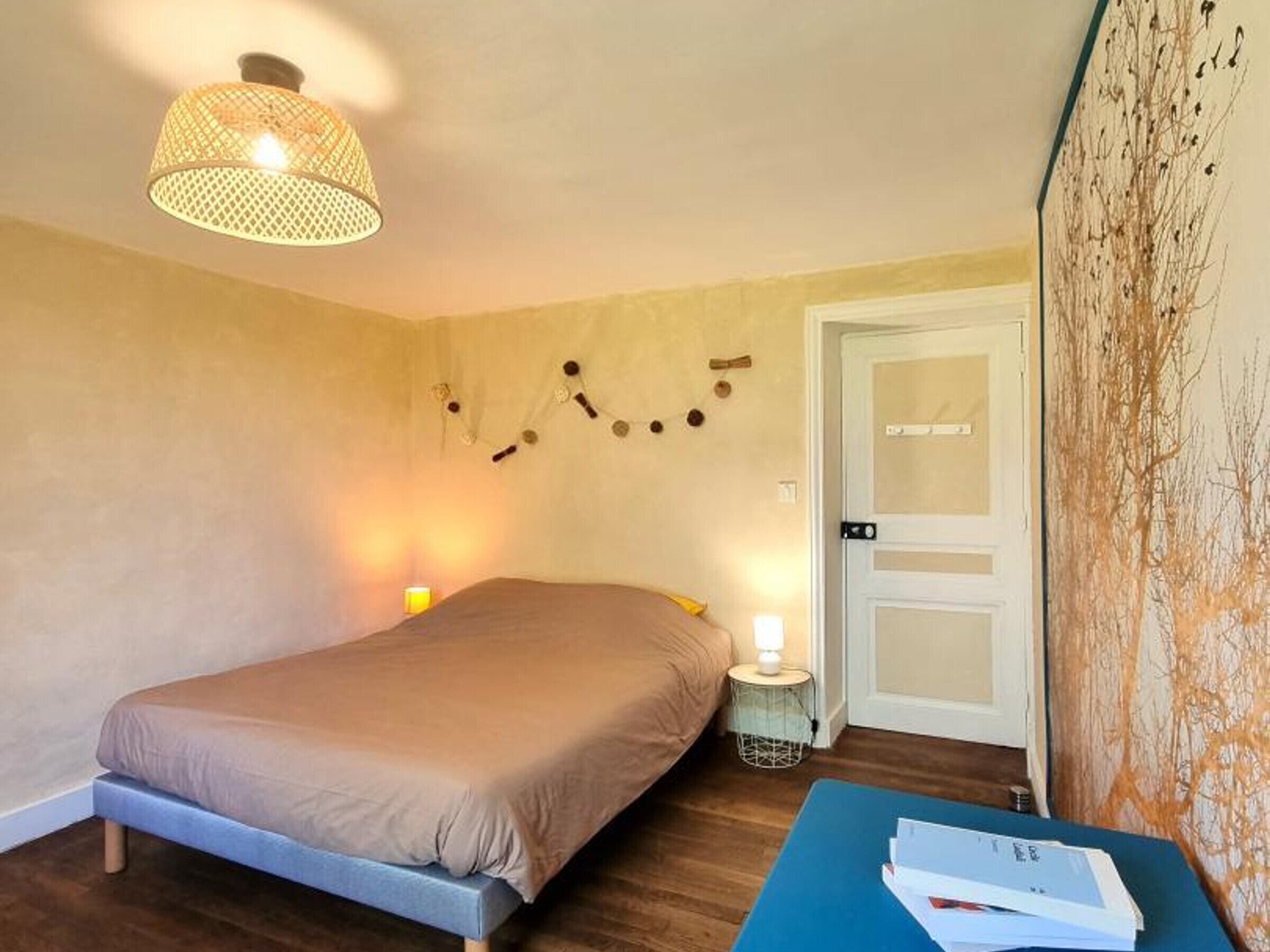 A bedroom with a bed, bedside lamps, a small table with papers, a door, and a wall-mounted light fixture.