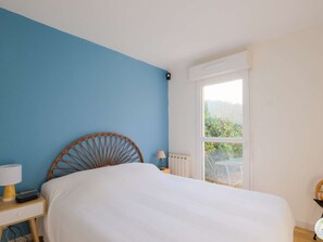 A bedroom with a bed, a nightstand, a lamp, a window, and a view of greenery outside. - Charming T3 Apt in Biscarrosse - Terrace, Garden, Near Lake, Sleeps 6 (Biscarrosse)