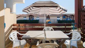 A patio with a table and chairs, a striped umbrella, and a building with blue doors in the background. - Biscarrosse Beach - T2 steps from the beach, private parking (Biscarrosse)