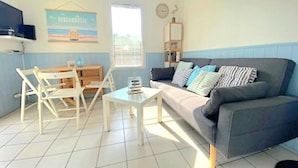 A living room with a sofa, a small table, a chair, a bookshelf, and a window. - Biscarrosse Beach Apt Terrace 4 pax with Parking (Biscarrosse)