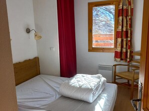 A single bed with a white mattress and pillows, a wooden headboard, a red curtain, a window with a view of trees, a wall-mounted lamp, and a wooden chair. - Cozy apartment, south terrace - 4 guests (Valloire)