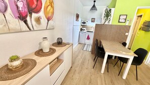 Une cuisine moderne avec des armoires blanches, un four à micro-ondes et un évier. On y trouve également un coin repas avec une table et des chaises, ainsi qu’un tableau représentant des fleurs accroché au mur.