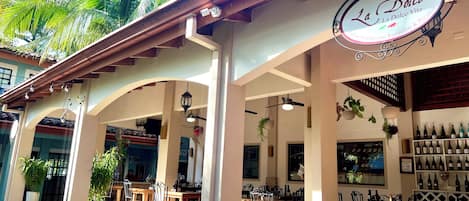 An outdoor dining area with tables and chairs, a sign for La Dolce Vita Restaurant & Pizzeria, and a wine rack with bottles.