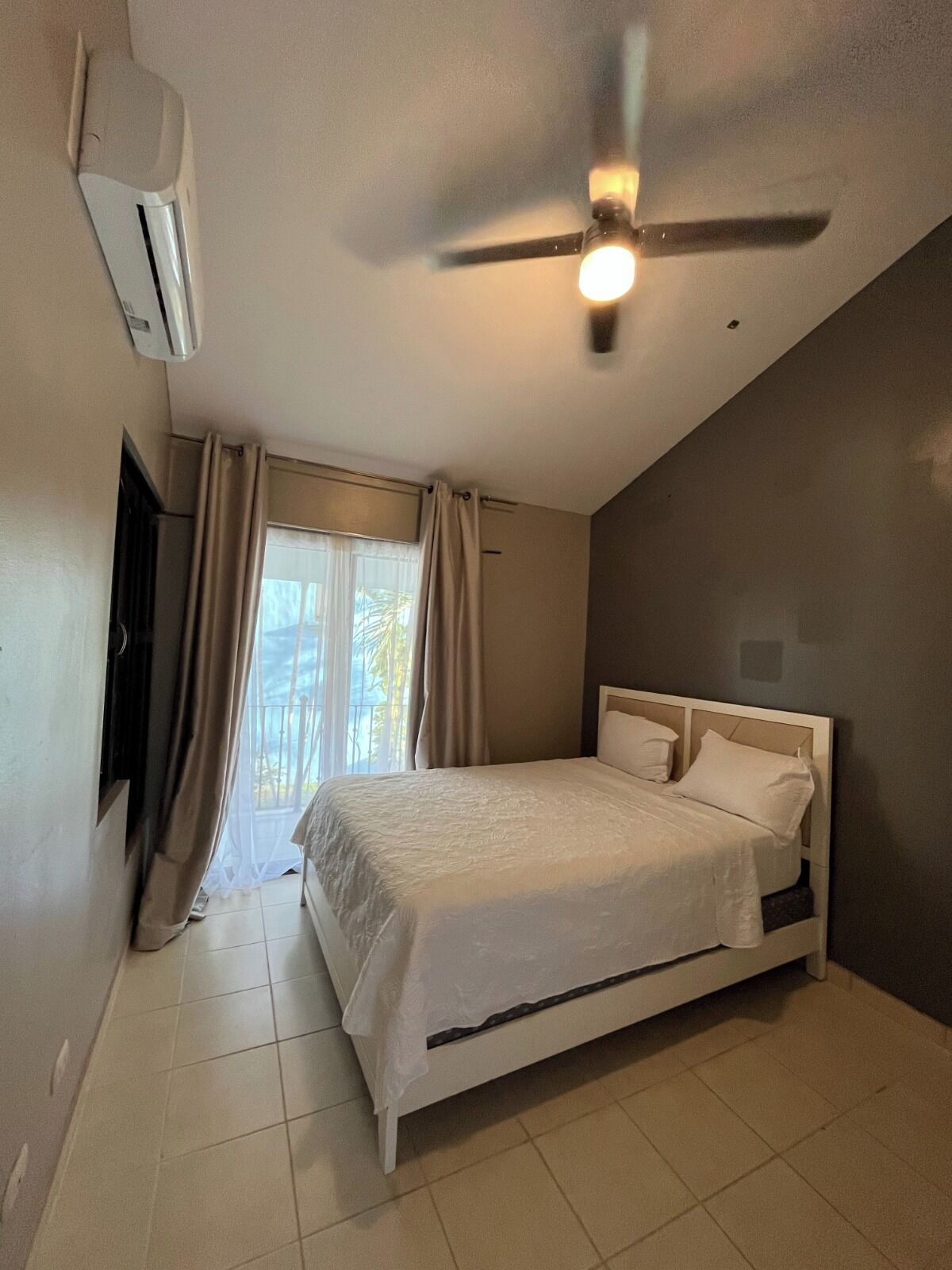 A bedroom with a bed, an air conditioning unit, a ceiling fan, and a window with curtains.