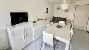 A modern kitchen with a flat-screen TV, white cabinets, a dining table with chairs, and decorative wall art. - Apartment with AC near beaches - 5 guests (Le Grau-du-Roi)