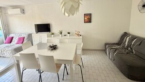 A modern living room with a white dining table, white chairs, a grey sofa, a flat-screen TV, and a ceiling fan. - Apartment with AC near beaches - 5 guests (Le Grau-du-Roi)