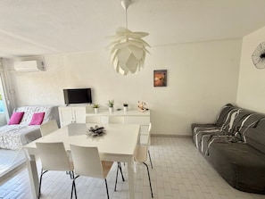 Ein modernes Wohnzimmer mit einem weißen Esstisch, weißen Stühlen, einem grauen Sofa, einem Flachbildfernseher und einem Deckenventilator.