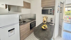 A compact kitchen with a refrigerator, sink, stove, microwave, and a countertop with a fruit bowl. - Apartment with AC near beaches - 5 guests (Le Grau-du-Roi)
