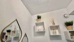A bathroom with white walls, a mirror with a gold frame, and shelves with plants and toiletries. - Apartment with AC near beaches - 5 guests (Le Grau-du-Roi)