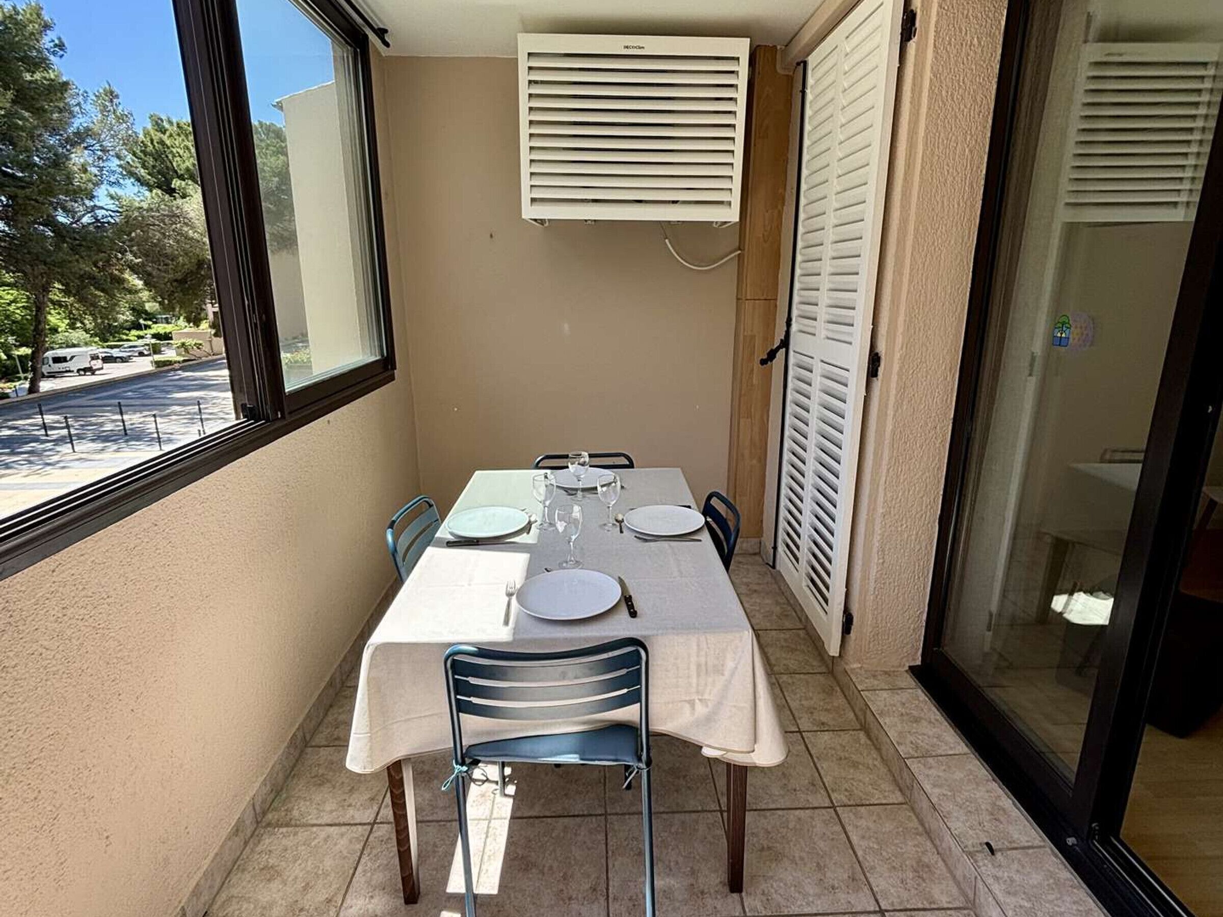 Un balcón con una mesa dispuesta para dos, una ventana y una puerta con contraventanas.