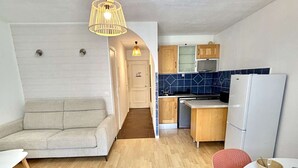 Een kleine leefruimte met een bank, een eettafel met een stoel, een keuken met kastjes en een koelkast, en een hanglamp.
