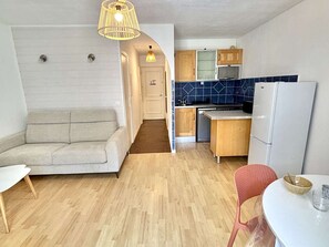 Ein kompakter Wohnbereich mit Sofa, Esstisch und Stuhl, Küchenzeile mit Schränken und Kühlschrank sowie einer Pendelleuchte.
