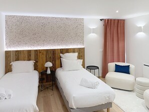Habitación de hotel con dos camas, un cabecero de madera, una mesita con una lámpara, una silla y un ventanal con cortinas.