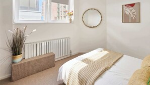 A bedroom with a bed, a window with a view of buildings, a radiator, a woven bench, and a wall-mounted mirror. - Boutique One Bed Apartment in Kemp Town (Brighton and Hove)