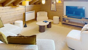 A cozy attic room with wooden beams, a white sofa, a round coffee table, a TV, and a large window overlooking a building. - Spacious apartment, Mont Pourri view - 8 guests (Montvalezan-La Rosière)