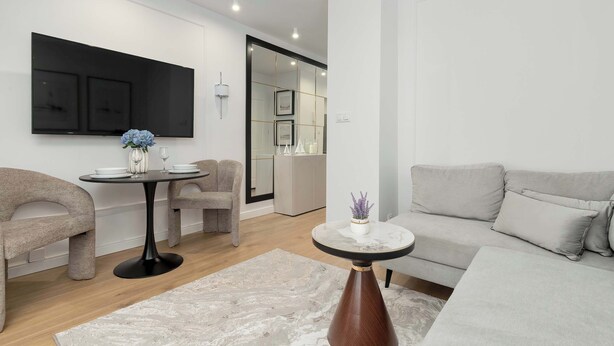 A modern living room with a sofa, a dining table with chairs, a flat-screen TV, and a large mirror.