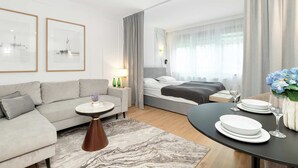 A modern hotel room with a sofa, a bed, a dining table with plates and glasses, and two framed pictures on the wall.