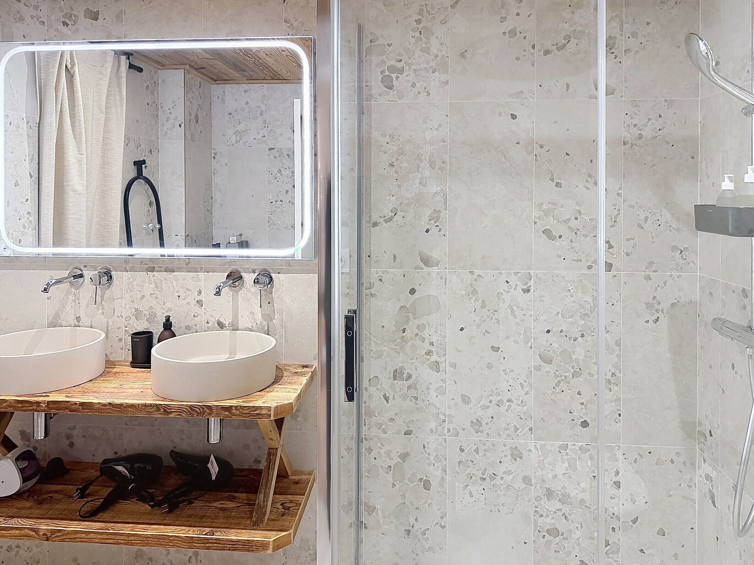 Un baño con dos lavamanos, ducha y espejo con estante de madera debajo.