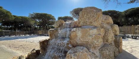 A natural rock formation with a cascading water feature, surrounded by a sandy area and trees.