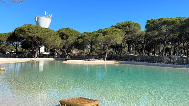 A clear pool with a wooden deck in the foreground, surrounded by trees and a white building with a tower in the background. - La Tamarissière Village  (Agde)