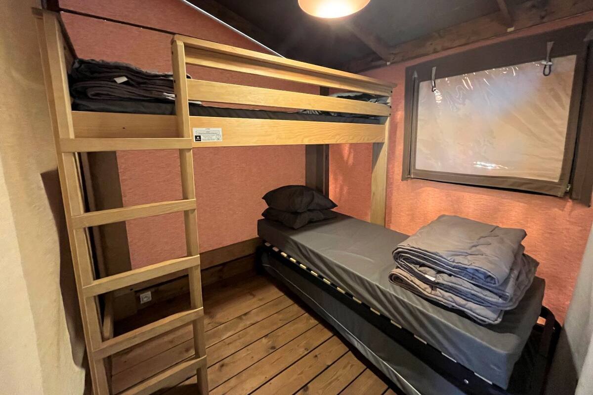 Una cama litera con escalera, una cama individual con colchón y almohadas, una ventana con persianas y un suelo de madera.