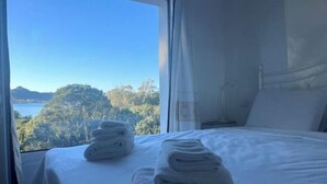 Quarto com cama, mesa de cabeceira e janela com vista para uma floresta e água.