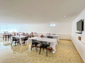 A spacious dining area with multiple tables and chairs, a flat-screen TV mounted on the wall, and a marble floor.