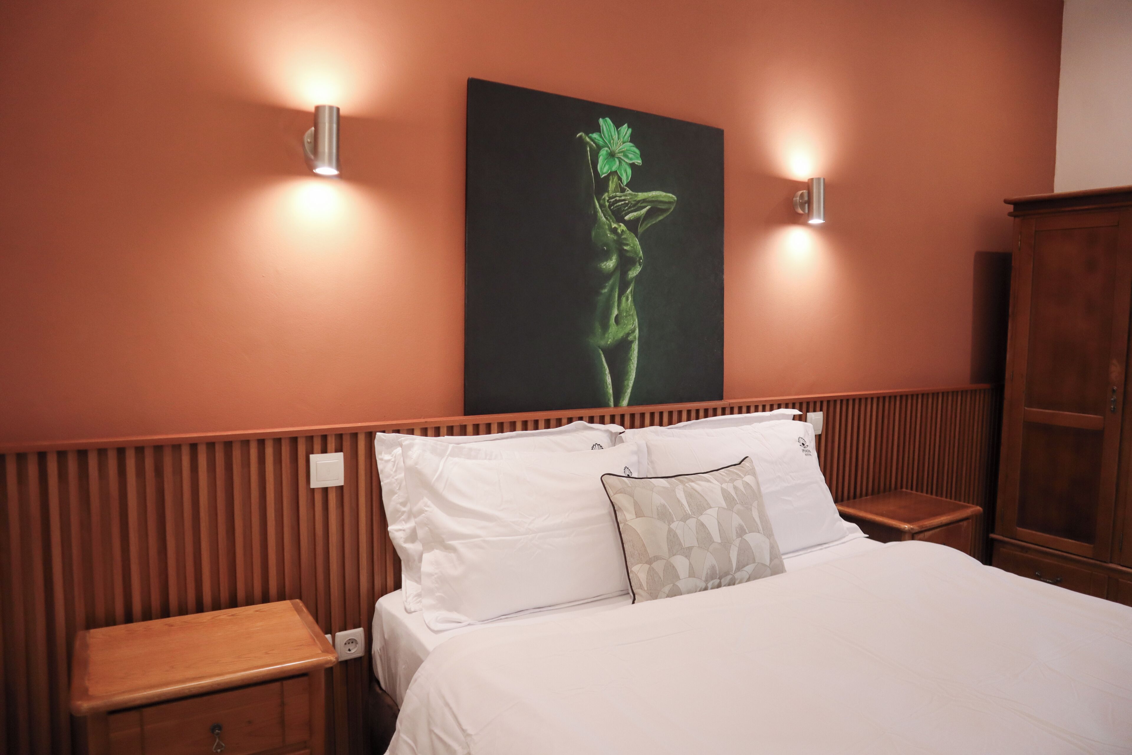 A hotel room with a bed, two bedside tables, a nightstand, a wooden cabinet, and a wall-mounted artwork.