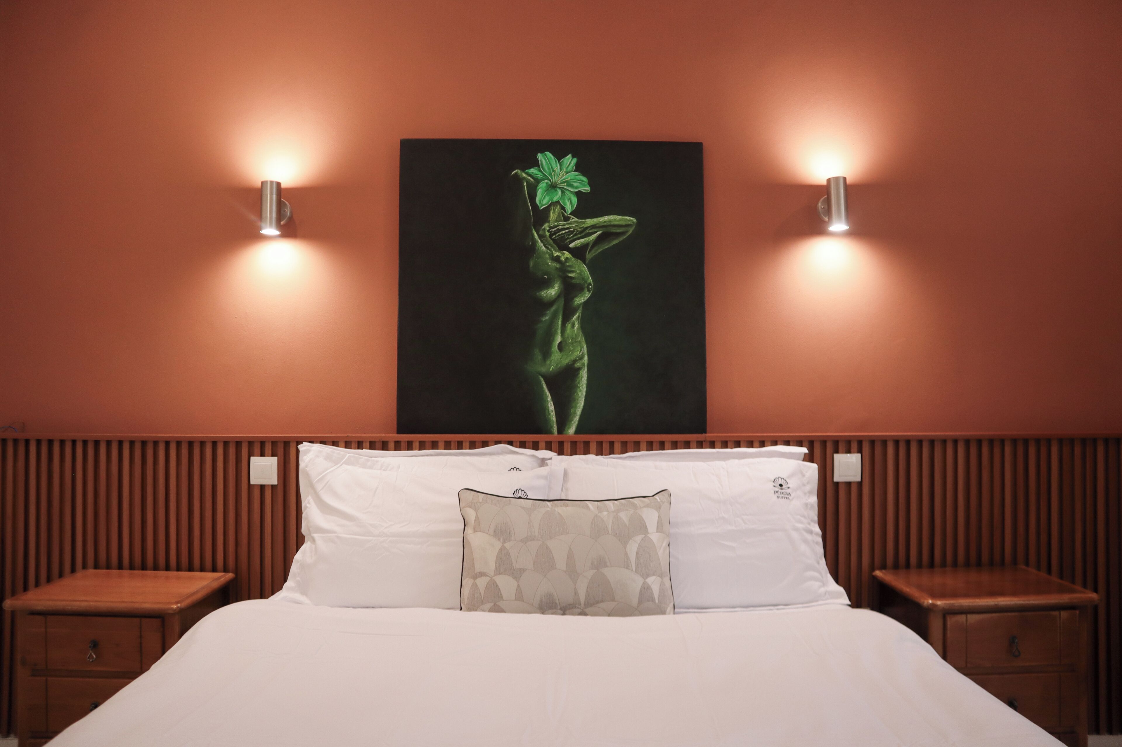 Quarto de hotel com cama, duas mesas de cabeceira, uma obra de arte na parede e duas arandelas.