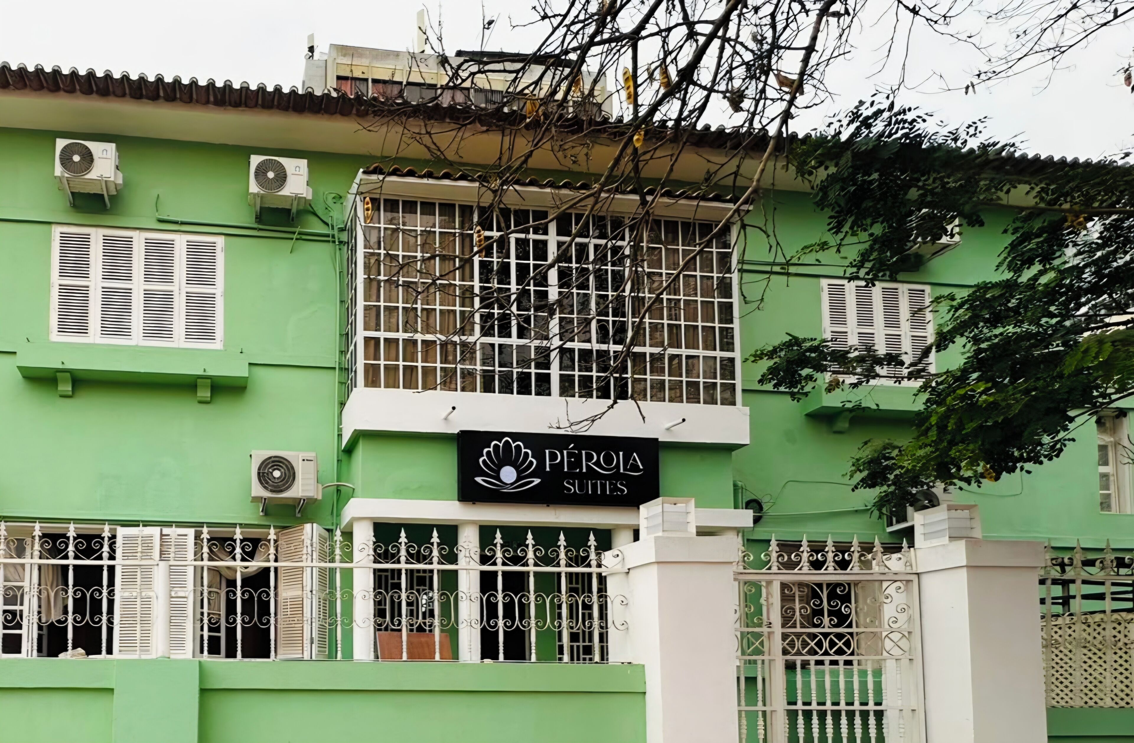 Un edificio con fachada verde, varios equipos de aire acondicionado, un letrero que dice 'Pérola Suites' y una entrada con portón.