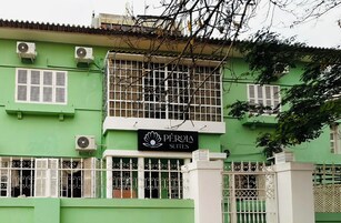 Um edifício com fachada verde, várias unidades de ar condicionado, uma placa com o nome 'Pérola Suites' e um portão de entrada.