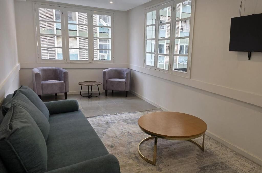 Een kamer met een groene bank, twee fauteuils, een klein rond tafeltje en grote ramen met jaloezieën.