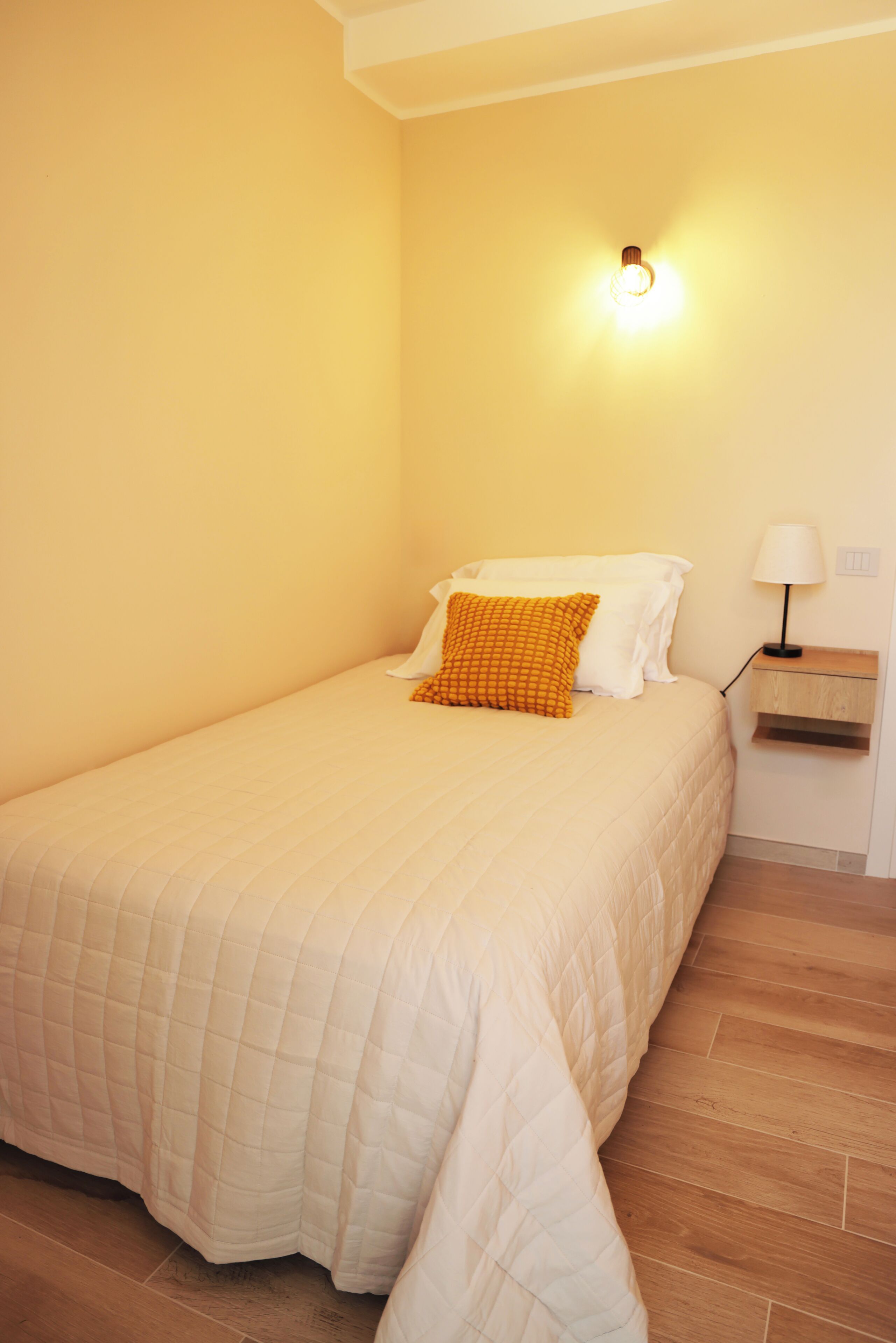 Uma cama de solteiro com colcha acolchoada, uma mesinha de cabeceira com abajur e luminária fixada na parede.