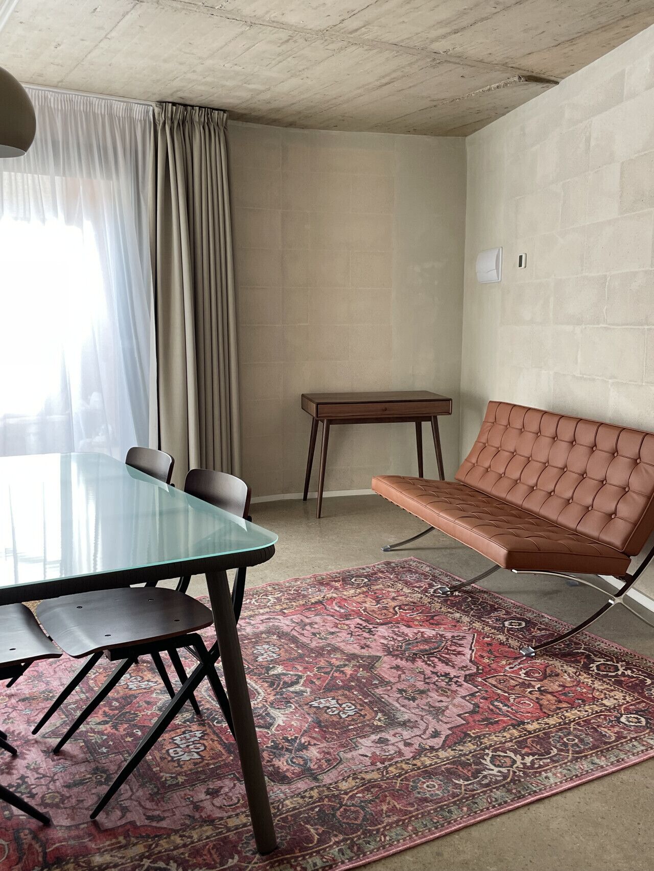 A room with a glass-top table, a brown leather sofa, a patterned rug, and a window with curtains.