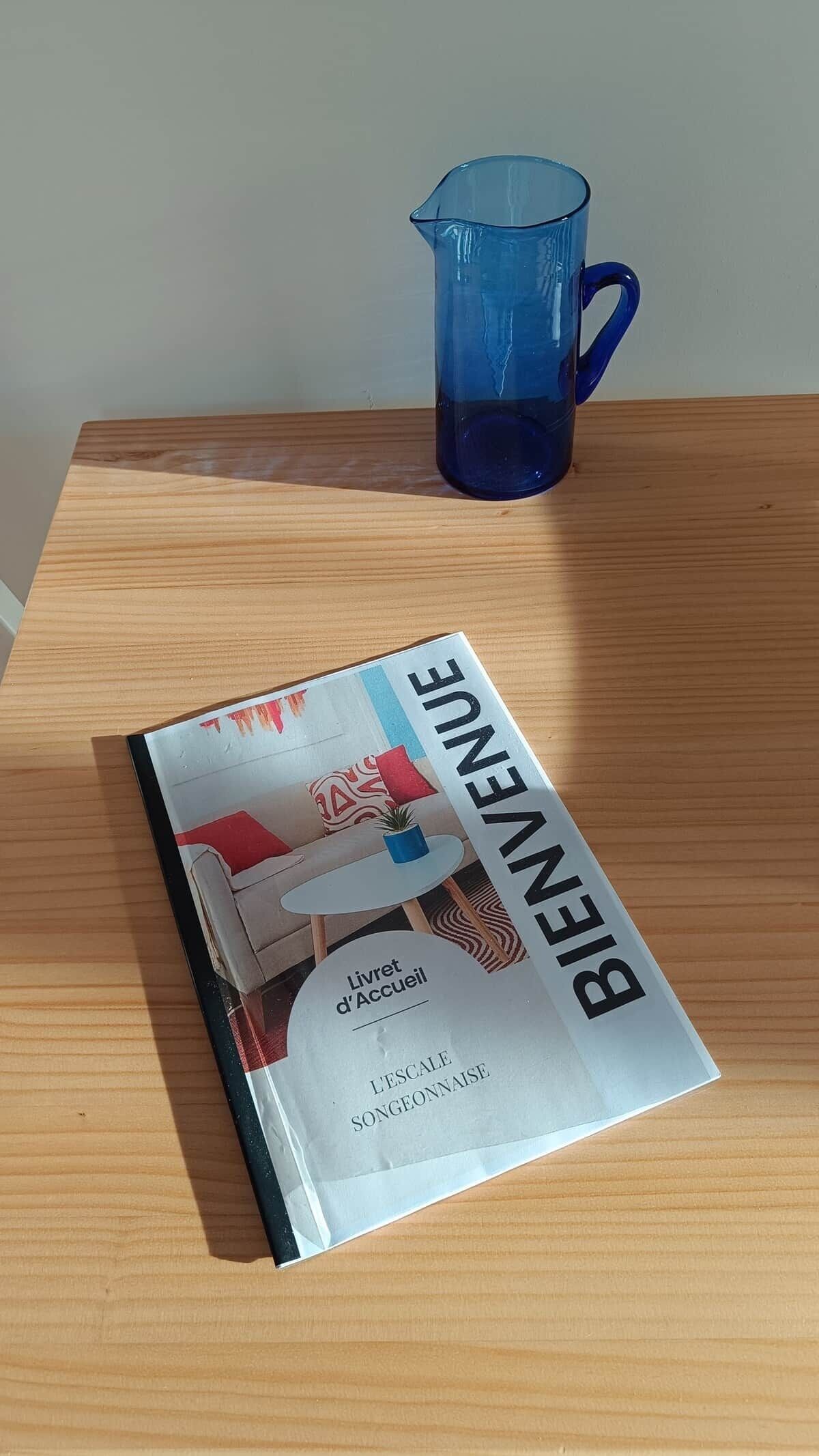 En bog med titlen 'Bienvenue' på et træbord med en blå glaskande ved siden af.